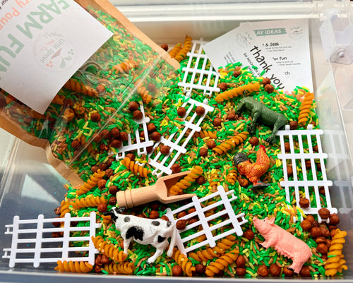 A clear plastic sensory bin filled with green and orange spiral pasta, white fence pieces, small brown beads, and farm-themed toy animals including a cow, pig, chicken, and horse. A brown paper bag labeled Farm Fun is tipped into the bin, along with printed cards, in a bright studio-style setup.