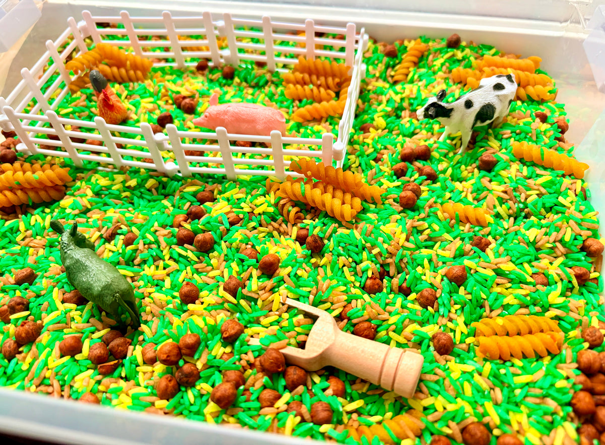 A sensory play tray filled with bright green and yellow rice, brown pellets, orange corn-shaped pieces, a white fence, a small pink pig, a cow figurine, a donkey figurine, and a wooden scoop.