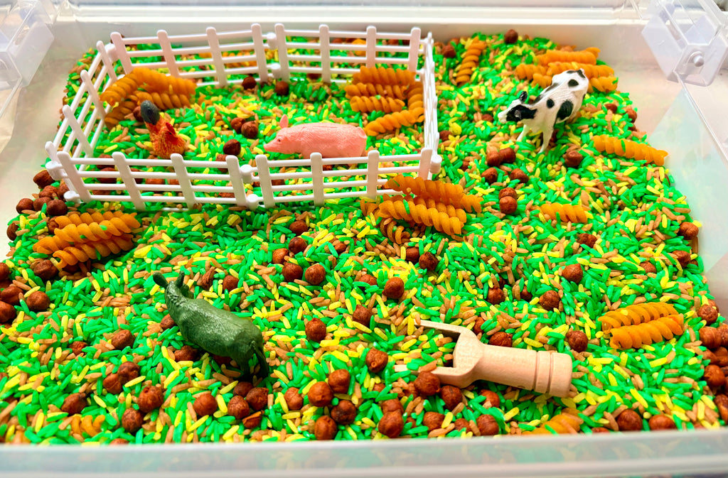 A children's sensory bin filled with bright green and yellow rice, brown pellets, yellow pasta resembling hay, a white plastic fence, a pink pig figure, a black-and-white cow figure.