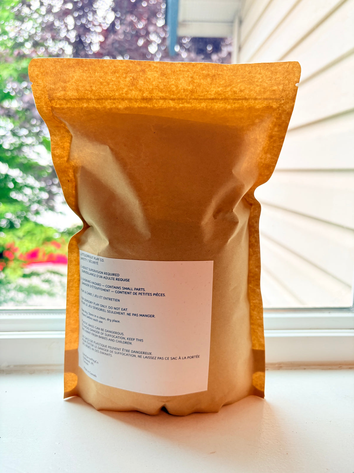 A stand-up kraft paper pouch with a white label, photographed on a windowsill with a window and outdoor greenery in the background. Showcasing safety label.